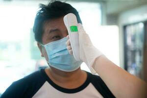 Asian waitress woman wearing face masks and holding an infrared forehead thermometer to check body temperature for virus symptoms of customers before entering the restaurant coffee shop. photo