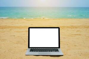 Laptop with blank screen for creative design on sand beach nearby blue sea background with sun ray effect. Computer notebook with monitor clipping path for present landing page design mock up template photo