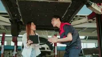 Asian female auto business entrepreneur discusses with male mechanical worker, checklist undercarriage of lifted electric car, fix and repair at maintenance garage, automotive service works industry. video