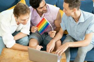 Triple young man sitting in the living room while using laptop to video call with rainbow flag on desk. LGBTQ people lifestyle and love emotion. LGBT social network. selective focus photo