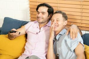 dos hombres en casual trajes sentado en sofá abrazando y acecho televisión con cerveza y Pizza en vivo habitación a hogar. lgbt gay relación concepto. foto