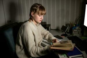 un sobrecargado de trabajo estudiante se sienta a noche con un libro. hacer notas en un cuaderno en el oscuro. foto