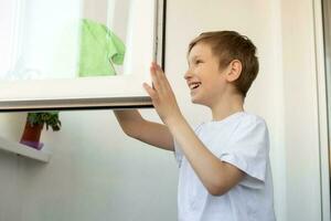 A cute boy is cleaning, washing an window on the balcony and laughing. Do wet cleaning with a rag photo