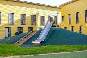 un metal diapositiva en un para niños patio de recreo con un verde césped. escalera a el colina. amarillo casa foto