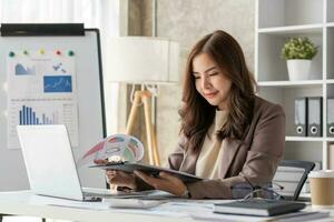 una alegre mujer de negocios que trabaja en una laptop en la oficina, una feliz y hermosa mujer de negocios asiática con traje formal trabaja en el lugar de trabajo. atractiva sonrisa de empleada de oficina. foto