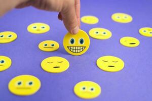 Hand holding yellow smiley face emoji between sad emojis on purple background photo