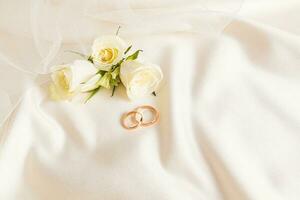 Two gold wedding rings on a beige satin background with live tea roses and part of the bride's veil. Chic layout for design. Wedding background. photo