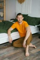 Young man is sitting at home on the floor near the bed and looking at the camera photo