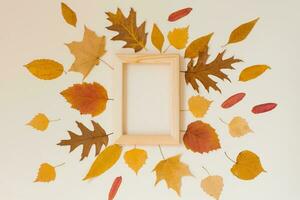 An empty wooden frame surrounded by yellow fallen leaves on a beige background. Autumn fla lay photo