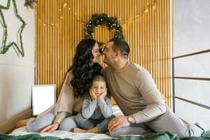 un joven familia con su hijo son sentado en el cama en el dormitorio, teniendo divertido y contento en el dormitorio decorado para Navidad foto