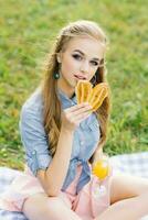 A beautiful young woman in a blue denim shirt and a pink skirt has a croissant in the garden at a picnic photo