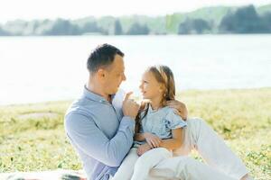 un contento padre sostiene su hija en su brazos. ellos tener divertido en naturaleza, sentado en un cobija por el lago en verano foto