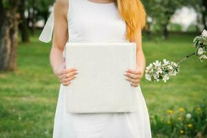 A photo book in a white leather cover in the hands of a woman in spring near a flowering branch of an apple tree