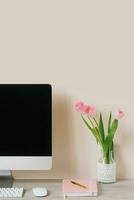 un computadora monitor, un ramo de flores de rosado tulipanes, y un cuaderno con un bolígrafo en un ligero de madera mesa en contra un beige pared. el lugar de trabajo de un persona de libre dedicación o estudiante foto