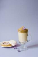 Beautiful dalgona drink a frothy coffee in a transparent mug and flowers of lilac on a purple background. Sweets on a plate, the donut and sweet marshmallows photo