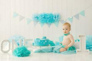 A one-year-old boy in blue shorts and a bow tie celebrates his birthday next to a cake on a stand and a decoration for his party photo