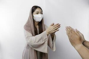 Portrait of a young beautiful Asian Muslim woman wearing a mask and headscarf gesturing Eid Mubarak greeting during pandemic photo