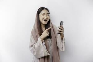 A happy Asian Muslim woman wearing a headscarf, holding her phone, isolated by white background photo