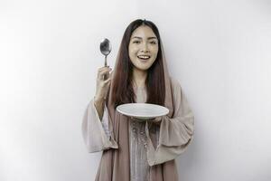 A smiling Asian Muslim woman is fasting and hungry and holding and pointing to a plate photo