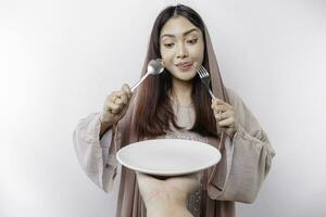 A smiling Asian Muslim woman is fasting and hungry and holding and pointing to a plate photo