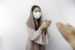 Portrait of a young beautiful Asian Muslim woman wearing a mask and headscarf gesturing Eid Mubarak greeting during pandemic photo