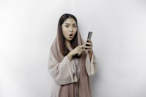 Shocked Asian woman wearing headscarf, holding her phone, isolated by white background photo