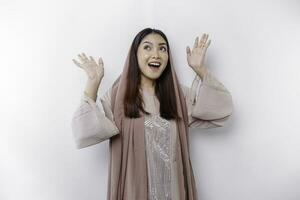 Excited Asian Muslim woman wearing headscarf pointing at the copy space above her, isolated by white background photo