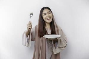 A smiling Asian Muslim woman is fasting and hungry and holding and pointing to a plate photo