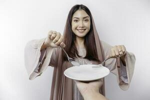 A smiling Asian Muslim woman is fasting and hungry and holding and pointing to a plate photo