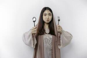 An Asian Muslim woman is fasting and hungry and holding cutlery while looking aside thinking about what to eat photo