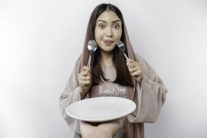 A smiling Asian Muslim woman is fasting and hungry and holding and pointing to a plate photo