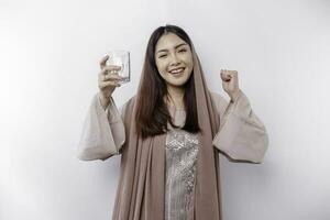 Joyful Asian Muslim woman wearing headscarf is drinking a glass of water, isolated on white background. photo