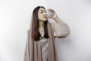 alegre asiático musulmán mujer vistiendo Pañuelo es Bebiendo un vaso de agua, aislado en blanco antecedentes. foto