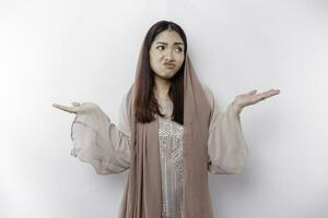 A thoughtful young Asian Muslim woman is wearing hijab and looks confused between choices, isolated by a white background photo