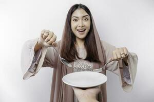 A smiling Asian Muslim woman is fasting and hungry and holding and pointing to a plate photo