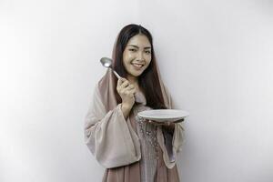 A smiling Asian Muslim woman is fasting and hungry and holding and pointing to a plate photo