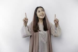 Excited Asian Muslim woman wearing headscarf pointing at the copy space above her, isolated by white background photo