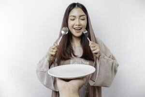 A smiling Asian Muslim woman is fasting and hungry and holding and pointing to a plate photo