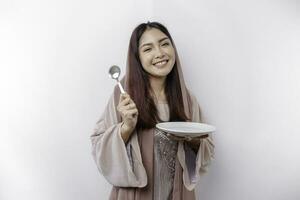 A smiling Asian Muslim woman is fasting and hungry and holding and pointing to a plate photo