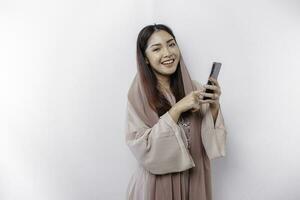 A happy Asian Muslim woman wearing a headscarf, holding her phone, isolated by white background photo