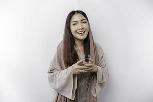 Joyful Asian Muslim woman wearing headscarf is drinking a glass of water, isolated on white background. photo