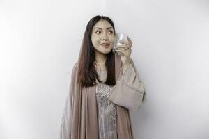 Joyful Asian Muslim woman wearing headscarf is drinking a glass of water, isolated on white background. photo