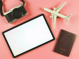 flat lay of digital tablet with blank white screen, airplane model, passport and digital camera isolated on pink background. travel planning concept. photo