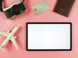 flat lay of digital tablet with blank white screen, airplane model, passport, credit card and digital camera isolated on pink background. travel planning concept. photo