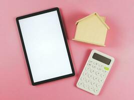 flat layout of digital tablet with white blank screen, wooden house model  and white calculator on pink  background with copy space, home purchase concept. photo