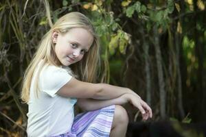 The little blonde girl sits near the green bushes and looks at the camera. photo