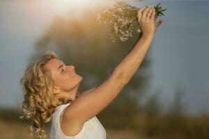 A beautiful middle-aged blonde with a bouquet of wildflowers on the background of the sun. photo