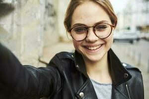 retrato de un contento mujer en lentes y en un cuero chaqueta al aire libre sonriente modelo foto