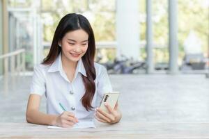 Young Asian woman student in uniform using smartphone and writing something about work.There are many documents on table her face with smiling working to search information for study. photo