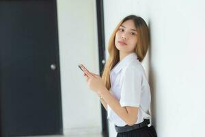 el retrato de una linda estudiante tailandesa asiática con uniforme sonríe alegremente mientras usa un teléfono inteligente en la universidad. foto